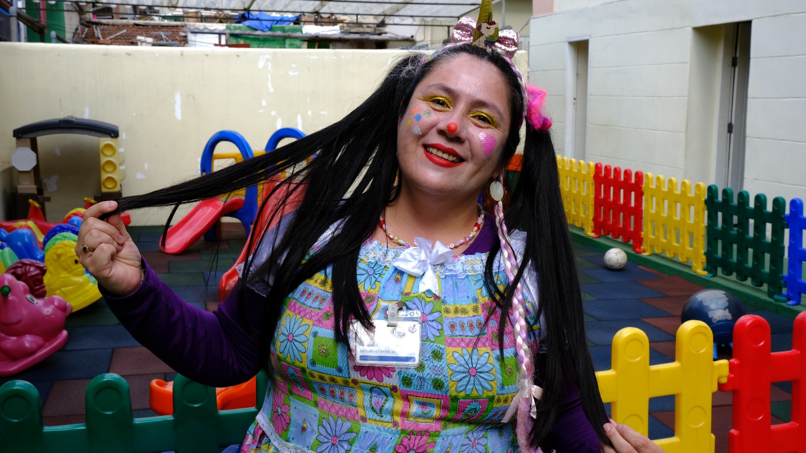 Na imagem,  a enfermeira Glorinha está pronta para mais uma ação, com roupas e maquiagem coloridas. O seu cabelo, comprido, liso e preto, tem duas traças, além de uma tiara de unicórnio e outros enfeites na cabeça. Ao fundo, há brinquedos infantis